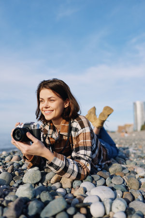 Female photographer lying on pebbles at the beach, smiling while holding a camera for outdoor...
