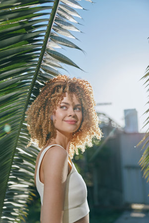 smiling woman with curly hair enjoys sunny outdoor setting near palm leaves, wearing white...