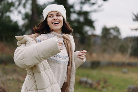 Woman outdoors wearing a winter coat, smiling, with scarf and beanie in a warm outdoor setting,...