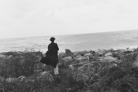 Solitary figure on a rocky coastline gazes toward the sea, a quiet moment of contemplation as the horizon meets the ocean in muted lightの写真素材