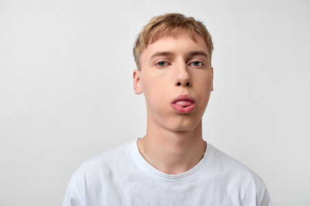 Young man with playful face sticking tongue out against a plain white background, casual wear and...