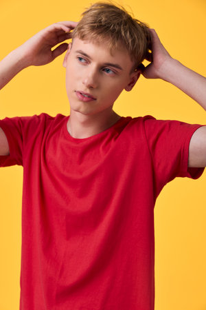 Young man in a red shirt, hands at his head, against a bright yellow backdrop, conveys a...