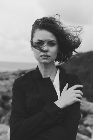 Woman on a windy coast in monochrome, wearing a jacket, hair blown, moody atmosphere, fashion...