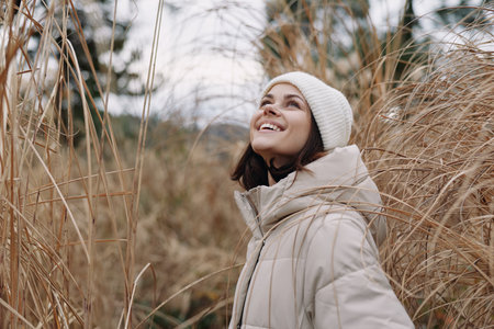 Woman in a warm coat and beanie outdoors, smiling among tall grasses in a chilly field, enjoying...