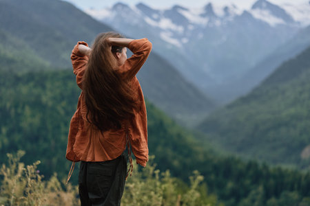 Woman stands outdoors with back to camera in a brown jacket, gazing at expansive mountains and lush...