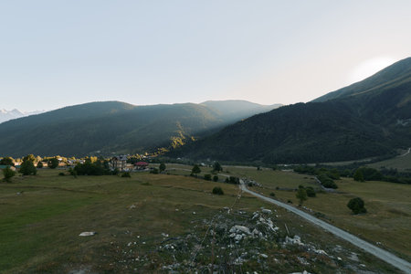 A tranquil mountain valley unfolds with a winding road, gentle fields, and distant hills bathed in...