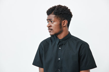 Young black man with dark skin wearing black shirt looking sideways on white background. Portrait of thoughtful African American male with short curly hair and casual style.の写真素材