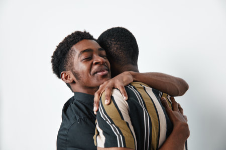 Two young black men hugging each other with eyes closed and smiles, showing friendship and affection in casual clothing on white background. Emotional connection and support.の写真素材