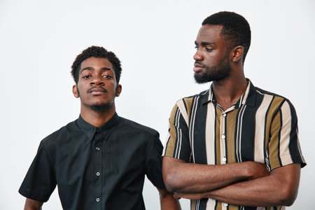 Two African American men with dark skin standing against white background, one wearing black shirt...