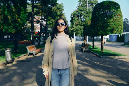 Woman in a long beige coat strolling through a sunny park, stylish sunglasses, casual chic mood,...