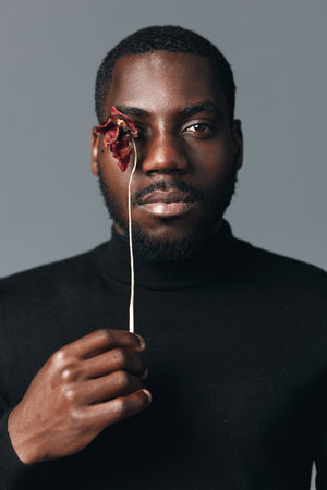 African man with dark skin holding dried flower in front of face wearing black turtleneck sweater...