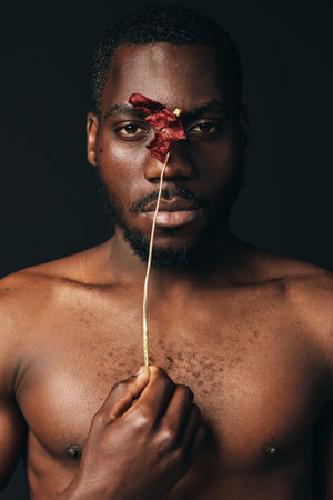 Black man with dark skin holding red flower in front of face in creative visual design style. Portrait with bare chest and artistic expression on dark background.の写真素材