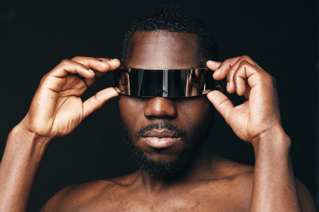 African man with dark skin wearing futuristic visor glasses in creative conceptual visual style. Portrait of confident male model with bare chest and modern accessory on black background.の写真素材