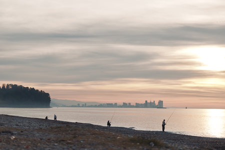Beach at dusk with calm water and silhouettes of anglers along the shoreline, warm sky and distant skyline, tranquil mood and gentle waves reflecting sunset light over pebbled shoreの写真素材