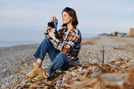 Photographer, woman with camera on a pebble beach, casual plaid shirt, jeans, hiking boots, capturing coastal scenery under clear sky, with wind in hair and calm ocean in the distance.の写真素材