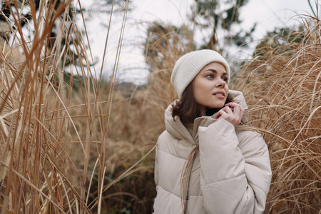 Winter outdoor portrait of a woman in a beige puffer jacket and a knit beanie among tall grasses, conveying calm weather and seasonal mood.の写真素材