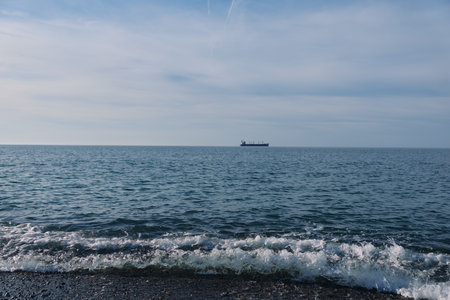 Sea and ocean scene with a distant ship on the horizon over calm water, gentle waves washing the shore, wide sky above, tranquil coastal view for travel and maritime explorationの写真素材