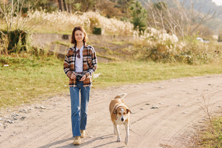 Woman walking with dog along a dirt path in a sunny outdoor setting, wearing a plaid shirt and jeans, capturing a peaceful nature moment on the trail.の写真素材