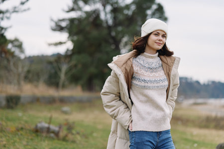 Woman in winter attire outdoors wearing a beige coat knitted sweater and white beanie standing in a...
