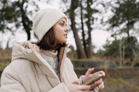 Woman in a beige winter coat and white beanie outdoors, looking to the side while using a phone, in a park with soft natural light.の写真素材
