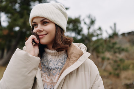 Woman talking on smartphone outdoors in winter, wearing a beige coat and knit hat, natural light,...