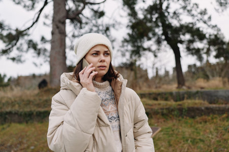 Woman in a light winter coat talks on a mobile phone outdoors in a park, surrounded by trees and open space, capturing a candid moment of remote communication in chilly weather.の写真素材