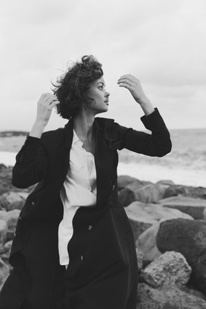 Woman in blazer and white shirt on a rocky coastline, wind in her hair, monochrome fashion portrait...