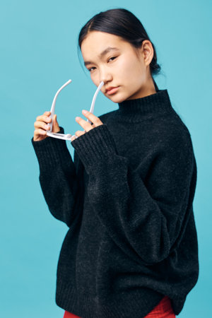 Young woman with black sweater putting on white sunglasses against blue background, posing with serious expression for fashion and style portrait photography in studio setting.の写真素材