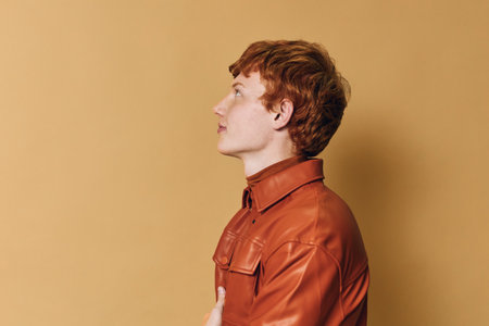 Profile of young man with red hair and freckles in brown leather jacket looking forward on beige background in studio. Side view portrait of thoughtful person with casual style and calm expression.の写真素材