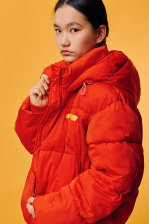 Young woman wearing red puffer jacket posing against yellow background with serious expression. Fashion portrait with warm clothing, winter style, and casual outfit for cold weather.の写真素材