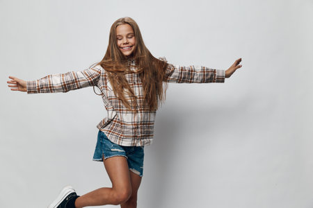 Girl jumping joyfully with eyes closed and arms outstretched wearing plaid shirt and denim shorts on white background with happy smile and long hair in motion.の写真素材