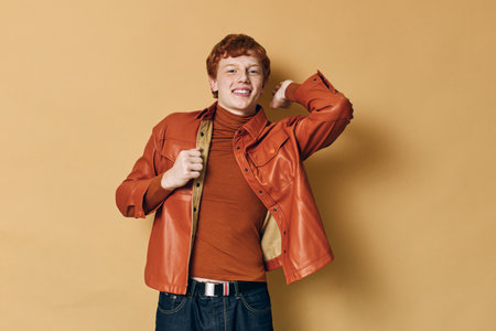 Young man with red hair wearing orange jacket and turtleneck poses smiling against beige background...