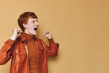 Young man with red hair celebrating success with clenched fists wearing brown jacket and turtleneck...