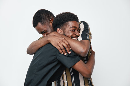 Two young black men with dark skin hugging each other with smiles on white background. Friendship,...
