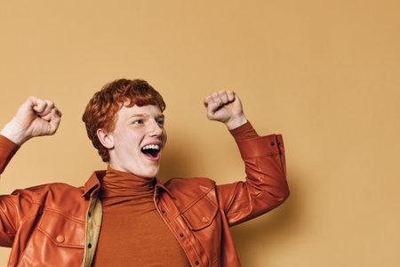 Young man with red hair wearing brown jacket and turtleneck celebrating with raised fists against beige background. Enthusiastic, happy and excited adult male portrait with expression of joy.の写真素材