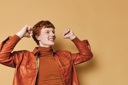 Young man with red hair wearing brown leather jacket and turtleneck celebrating with raised fists and smiling against beige background in studio. Cheerful emotional expression.の写真素材