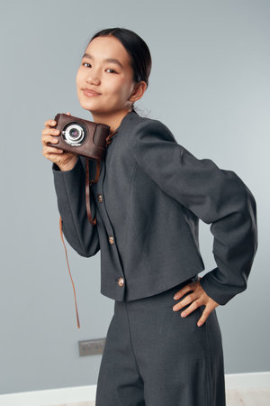 Young woman with short dark hair holding vintage camera smiling and posing in stylish gray suit on...