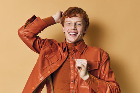 Young man with red hair smiling while wearing brown leather jacket and turtleneck shirt against beige background in studio portraitの写真素材
