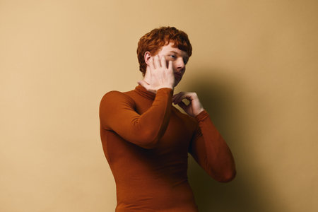 Young man with red hair in brown sweater talking on smartphone against beige background, casual communication and technology connection concept with thoughtful expression.の写真素材