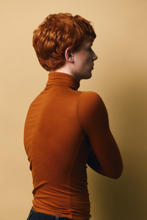 Portrait of young person with red hair and fair skin wearing orange turtleneck, standing sideways...
