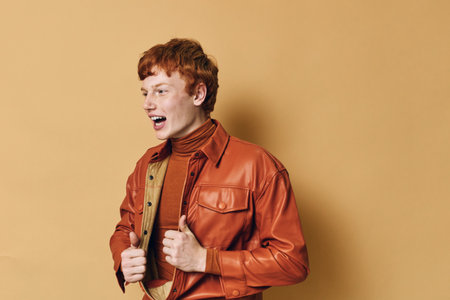 Young man with red hair wearing a brown turtleneck and orange leather jacket posing smiling with open mouth against beige background in studio. Casual fashion style portrait.の写真素材