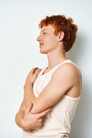 Young man with red hair and freckles wearing white tank top standing in profile against plain background with relaxed expression and folded arms.の写真素材