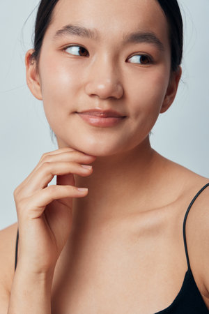 Young woman with bare shoulders and natural makeup looks sideways with gentle smile and hand near face against plain background, portrait of serene beauty and confidence.の写真素材