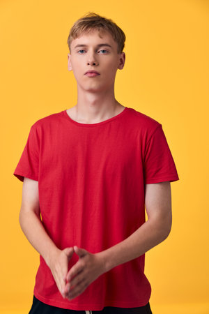 Red shirted young man stands in a studio against a bright yellow backdrop, hands clasped in front, neutral expression, bold minimal shot ideal for fashion, lifestyle or advertising useの写真素材