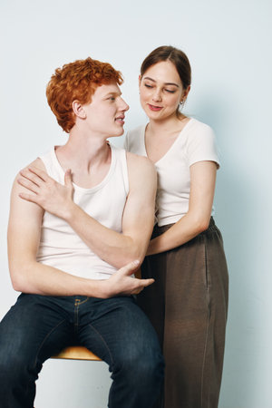 Young couple with man sitting on stool and woman standing behind him, showing affection and closeness in casual clothes on light background.の写真素材