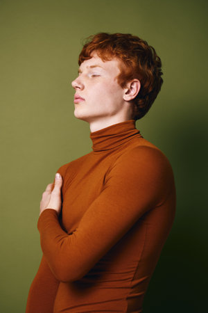 Portrait of young man with red hair wearing brown turtleneck looking sideways with closed eyes against green background. Fashion model posing with thoughtful expression in studio.の写真素材