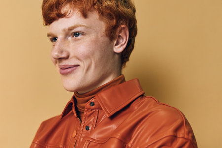 Young man with red hair and freckles wearing orange leather jacket and turtleneck smiling with confidence against beige studio background. Portrait of casual style.の写真素材
