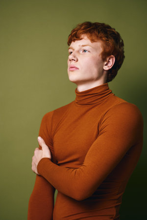 Young man with red hair and fair skin posing confidently in brown turtleneck sweater against green...