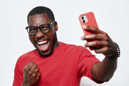 African American man with dark skin and glasses taking selfie with smartphone, smiling and celebrating success in casual red shirt on white background.の写真素材