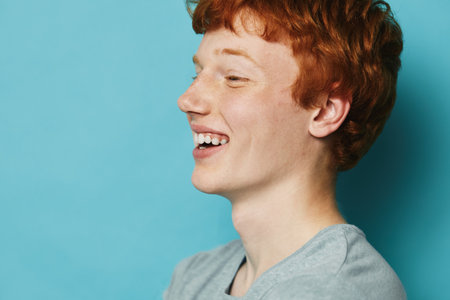 Teenager boy with red hair and freckles smiling sideways against blue background. Casual portrait...